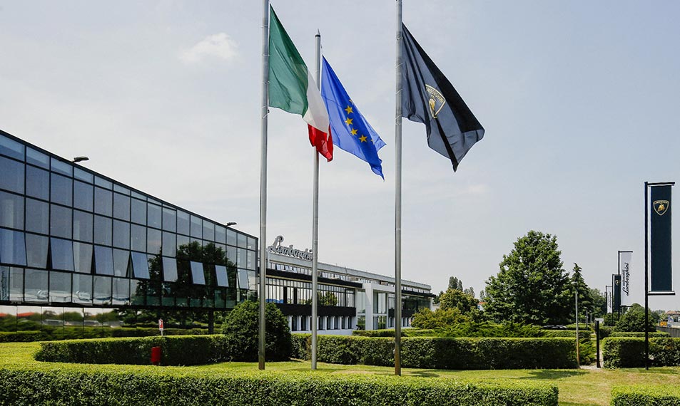 The image shows the Lamborghini corporate building, three flags flying before it. Starting from the left, these are the flags of Italy, Europe and a dark one bearing the Lamborghini crest.